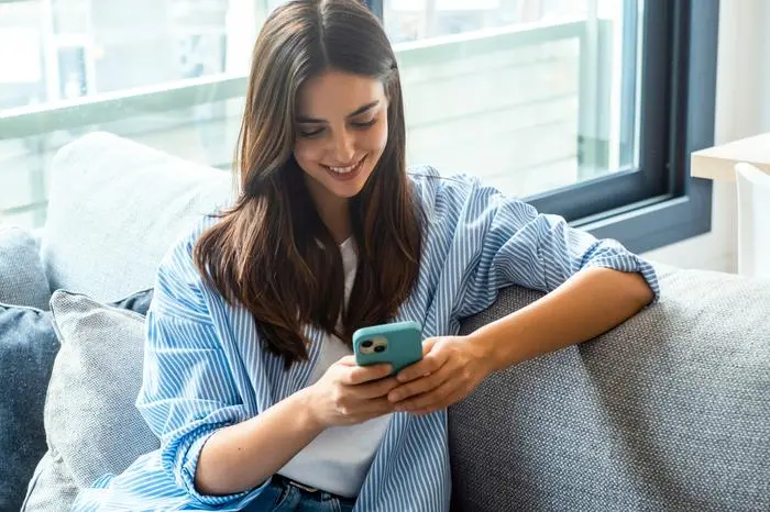 Happy girl checking social media while holding smartphone at home. Smiling young brunette woman using mobile phone app to play games, shop online, order delivery while relaxing on sofa. Scrolling.