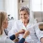 A young health visitor helping a happy sick senior woman sitting on bed at home.