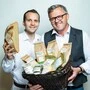 David und Johannes Tomic vom Biohof Tomic stehen nebeneinander. Sohn David hält ein halbes Brot in der Hand, Vater Johannes einen geflochtenen Korb mit Mehlen und Müsli darin