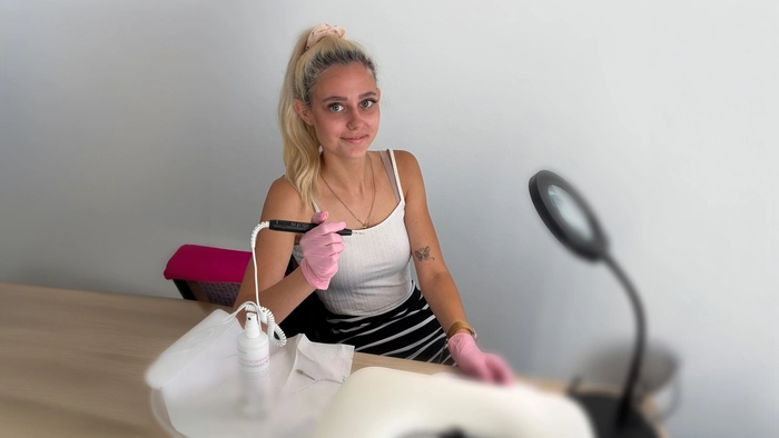 Nageldesignerin Leonie Stückelberger sitzt in ihrem Studio auf einem Stuhl. Sie trägt rosa Hygienehandschuhe und hält eine elektrische Nagelfeile in der Hand