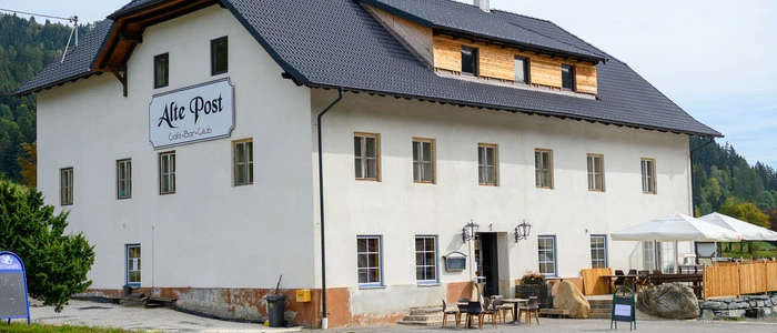 Das Bild zeigt eine Außenansicht des Gasthauses „Alte Post“ mit dem Sitzgarten neben dem Eingang. Dach und Fenster im Erdgeschoss sind neu, das Gebäude selbst ist sehr alt.