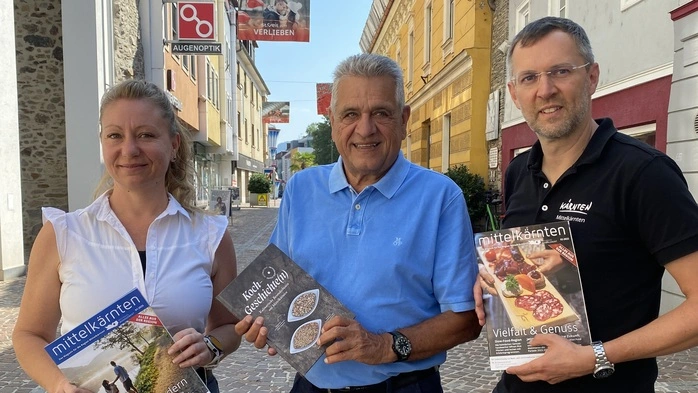Leader-Managerin Barbara Hudelist, Regionsobmann Gerhard Mock und Regionalmanagement-Geschäftsführer Gunter Brandstätter stehen nebeneinander am St. Veiter Hauptplatz. Sie halten das Regionsmagazin und das Kochbuch (beides Leader-Projekte) in Händen