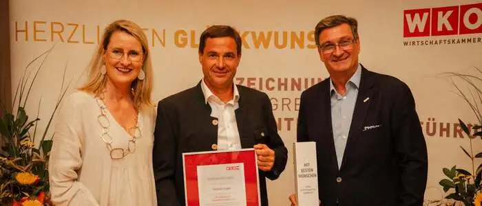 WK-Bezirksobfrau Eva Hoffmann, Unternehmer Wolfram Kogler, WKK-Präsident Jürgen Mandl stehen in einer Reihe, Kogler hält eine Ehrenurkunde in den Händen