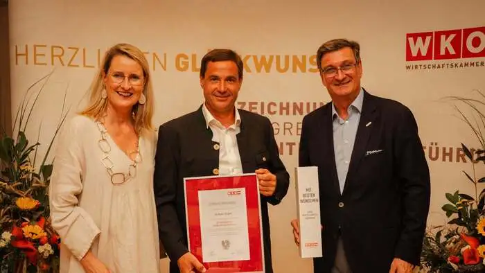 WK-Bezirksobfrau Eva Hoffmann, Unternehmer Wolfram Kogler, WKK-Präsident Jürgen Mandl stehen in einer Reihe, Kogler hält eine Ehrenurkunde in den Händen