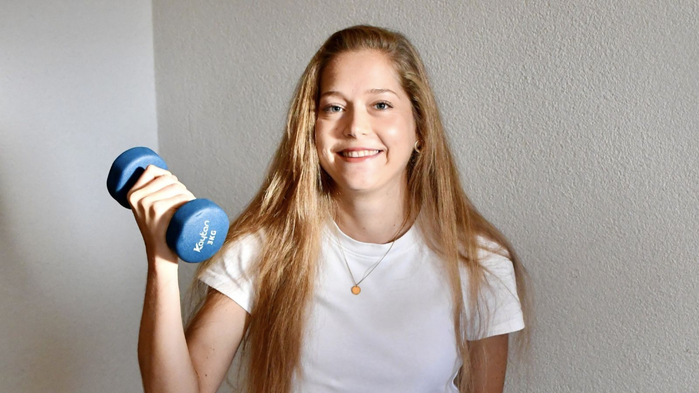 Physiotherapeutin Julia Jernej sitzt auf einem Gymnastikball und hält eine Hantel in der Hand. Sie trägt eine weiße Hose und ein weißes T-Shirt und hat lange hellbraune Haare.
