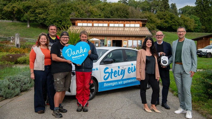 Vertreter der Diakonie de la Tour und des Unternehmens Go-e stehen vor einem Elektroauto und halten eine Wallbox (Ladestation) in der Hand.