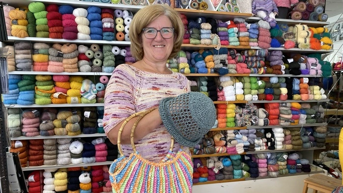 Unternehmerin Elisabeth Münzer-Kuess steht in ihrem Geschäft und hält einen gehäkelten Hut und eine Tasche in der Hand. Sie steht vor einem Regal voller bunter Wollknäule