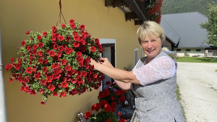 Barbara Wildpanner steht bei ihrem Haus und zupft bei einem der hängenden Blumentröge an den Blumen. Sie hat kurze blonde Haare, trägt ein Trachtengilet und eine passende Bluse und lächelt in die Kamera.