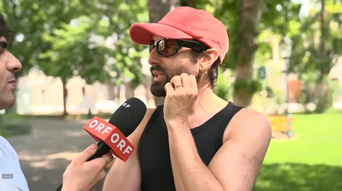 Tom Neuwirth mit roter Schirmkappe vor einem ORF-Mikro. Am Ringfinger seiner linken Hand ist ein goldener Ring zu sehen