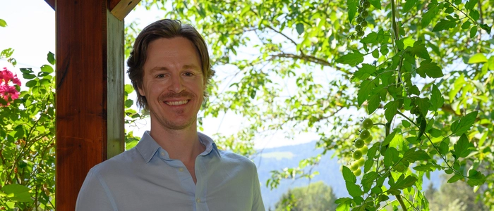 Lied- und Opernsänger Georg Klimbacher steht auf der Terrasse seines Elternhauses mit Blick ins Grüne. Er trägt ein blaues lockeres Leinenhemd: