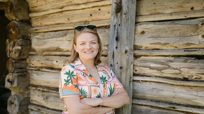 Eine Frau in einer Bluse mit Blumenmuster lehnt an der Wand einer Holzhütte und lächelt.