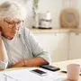 People, age, technology and finances. Depressed unhappy retired woman paying domestic bills online, trying hard to make both ends meet, sitting at kitchen table, surrounded with papers, using gadgets