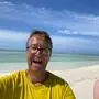 Ein Mann in einem gelben T-Shirt steht auf einem Strand und lacht in die Kamera | Reinhard Krämmer auf „seiner“ Insel Kaaddoo im Indischen Ozean