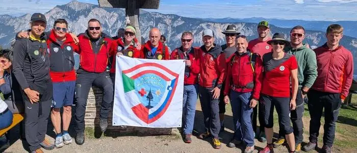 Eine Gruppe Bergretter und Bergretterinnen vor einem Gipfelkreuz mit einer Fahne, im Hintergrund Bergpanorama