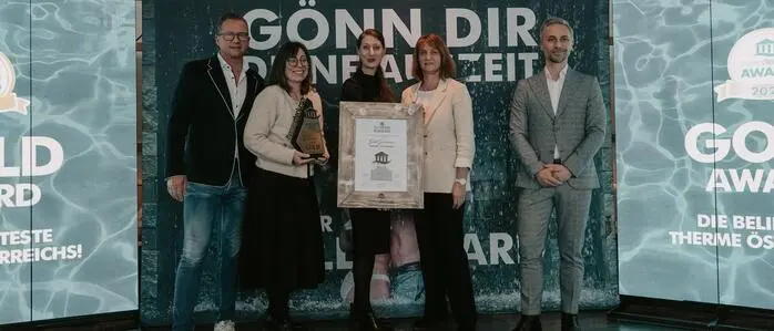 Man sieht drei Frauen und zwei Männer vor einer Plakatwand, die Frau in der Mitte hält eine große Auszeichnung in Händen | Das Rogner Bad Blumau wurde als beste Therme Österreichs ausgezeichnet