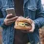 Das Bild zeigt eine Person, die einen Burger und ein Smartphone in der Hand hält