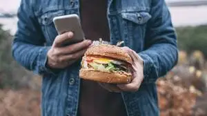 Das Bild zeigt eine Person, die einen Burger und ein Smartphone in der Hand hält