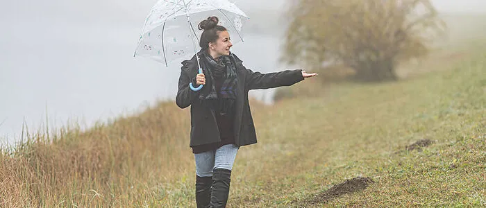 Das Bild zeigt eine Frau mit einem Regenschirm, die vor einem nebeligen Hintergrund steht
