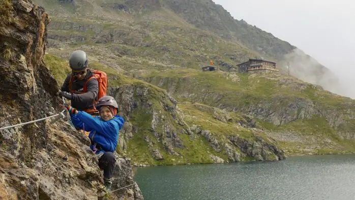 Ein Kind klettert auf einem Klettersteig. Hinter dem Kind ist ein Betreuer