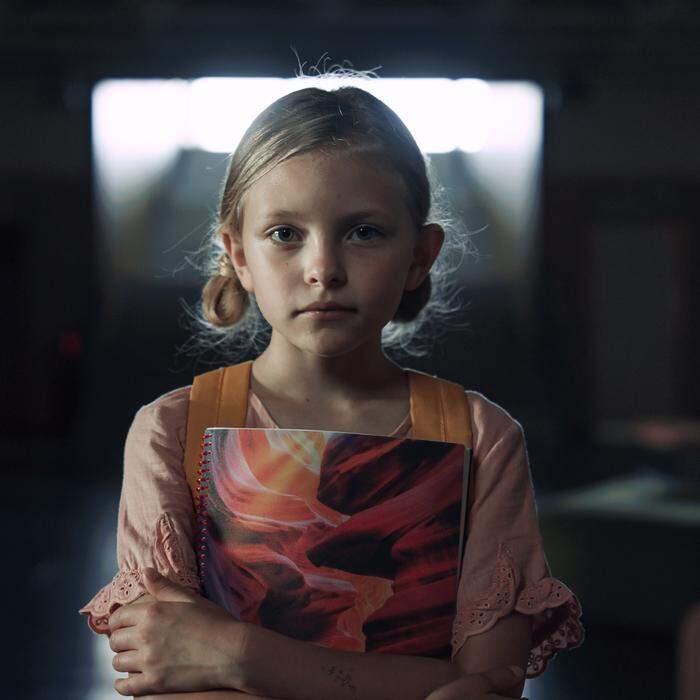 Portrait of upset frightened schoolgirl standing dark hallway alone. Lonely depressed elementary age girl posing in school corridor with books. Helpless blonde child full of despair looking camera.