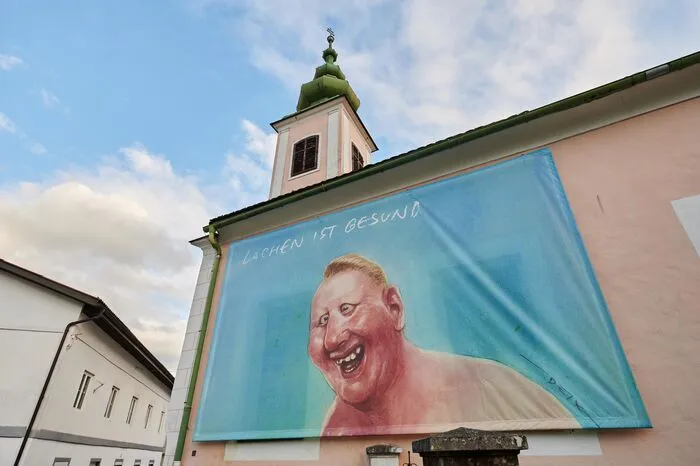 Großflächige Fassadengestaltungen in der Innenstadt von Bleiburg mit Motiven von Manfred Deix und WernerBerg erweitern die Ausstellung in den öffentlichen Raum.