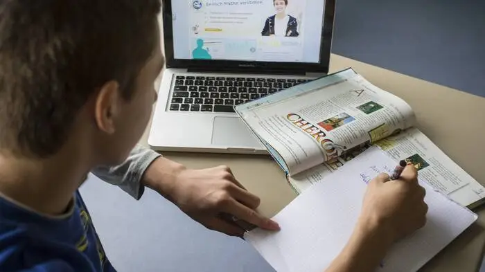 Bild zeigt schreibenden Schüler mit Schulbuch vor dem Laptop