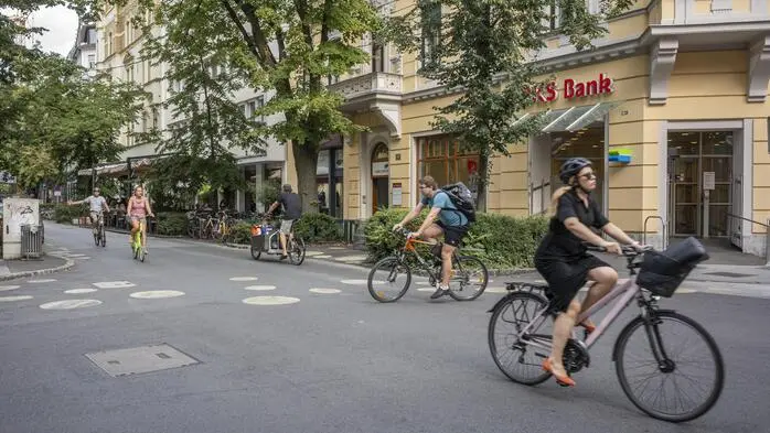 Große Bäume in der Kaiserfeldgasse im Hintergrund, davor Radfahrer, die die Straße queren