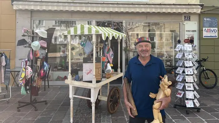Ein Mann hält eine selbstgemachte Pfeffermühle und einen selbstgemachten Pinocchio in den Händen. Er steht vor einem Geschäft und lächelt in die Kamera.