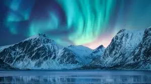 Aurora borealis over the snowy mountains, coast of the lake and reflection in water. Northern lights above snow covered rocks. Winter landscape with polar lights, fjord. Starry sky with bright aurora