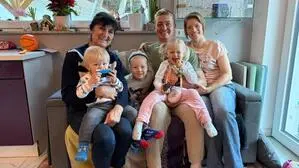 Zwei Frauen, jeweils rechts und links außen, in der Mitte ein Mann und drei Kinder sitzen auf einer Couch und lachen in die Kamera | Mama Zalika, Matija und seine Frau Maja Smrtnik mit den drei Kindern