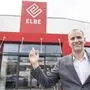 Ein Mann steht vor einem Geschäft mit der Aufschrift „Elbe“ und zeigt mit dem rechten Zeigefinger auf das rote Logo über der Eingangstür