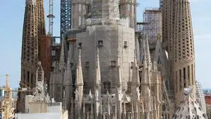 Das Bild zeigt die berühmte Basilika <strong>Sagrada Família</strong> in Barcelona, Spanien. Das monumentale Bauwerk befindet sich weiterhin im Bau, was durch die zahlreichen Baugerüste und Kräne deutlich wird. Im Vordergrund sieht man Wohngebäude, während sich dahinter die eindrucksvollen, reich verzierten Türme der Basilika in den Himmel erheben. Die markante Mischung aus gotischen und modernistischen Elementen sowie die aufwendige Fassadengestaltung sind typisch für den Architekten Antoni Gaudí. | Seit 1882 wird an Gaudís Meisterwerk gearbeitet. Nun ist ein Ende in Sicht