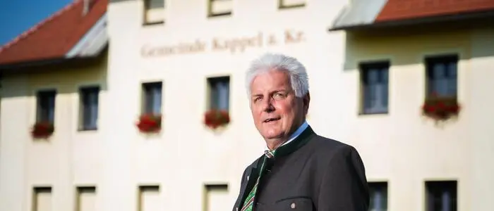 Ein Mann mit grauen Haaren steht vor einem gelblichen Gebäude.