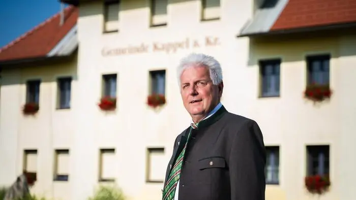 Ein Mann mit grauen Haaren steht vor einem gelblichen Gebäude.