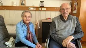 Eine ältere Frau sitzt auf einem Stuhl, neben ihr sitzt ihr Mann in einem Rollstuhl, beide lächeln in die Kamera