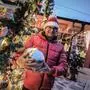 Ein Mann mit roter Weihnachtsmütze hält eine große Weihnachtskugel. Im Hintergrund sieht man ein Haus voller Lichterketten | Bei Volker Jürgen Hartl ist Weihnachten tief im Herzen verankert