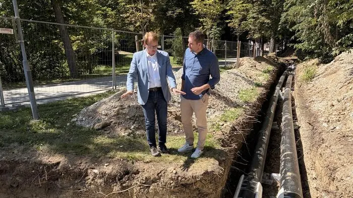 Zwei Fernwärmeleitungen im Boden sind zu sehen, die einen 90-Grad-Knick machen. Daneben steht Bioenergie-Geschäftsführer Jakob Edler mit weißem Hemd und hellem Sakko, der gerade dem Bürgermeister von Premstätten Matthias Pokorn (ÖVP) etwas erklärt