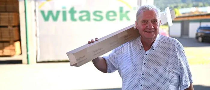 Ein Mann trägt eine Baumschutzhülle über der Schulter und lächelt in die Kamera. Im Hintergrund sieht man den Schriftzug „Witasek“