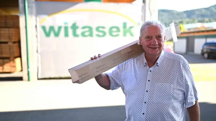 Ein Mann trägt eine Baumschutzhülle über der Schulter und lächelt in die Kamera. Im Hintergrund sieht man den Schriftzug „Witasek“