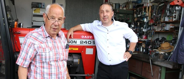 Zwei Männer stehen in einer Werkstatt. Ein Mann im weißen Hemd lehnt sich an einen Traktor