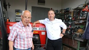 Zwei Männer stehen in einer Werkstatt. Ein Mann im weißen Hemd lehnt sich an einen Traktor 