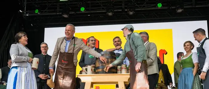 Mehrere Personen stehen auf einer Bühne. Landeshauptmann Mario Kunasek steht neben einem Bierfass und hat einen Holzhammer in der Hand.