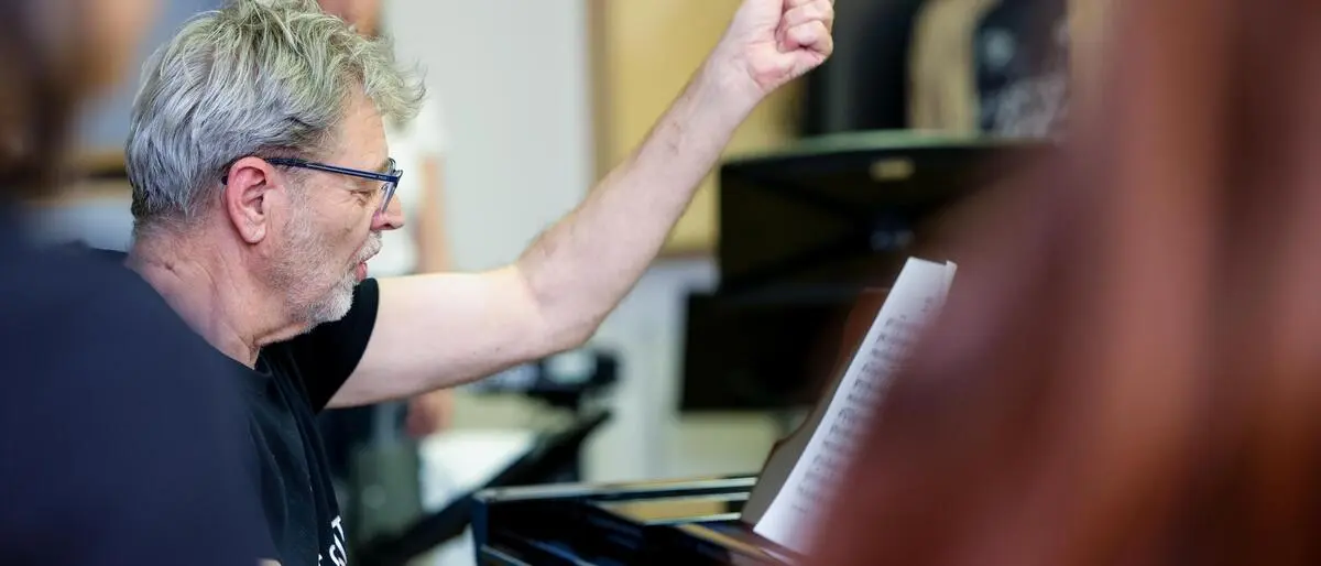 Mann sitzt am Klavier, die rechte Hand an den Tasten, mit der linken dirigiert er