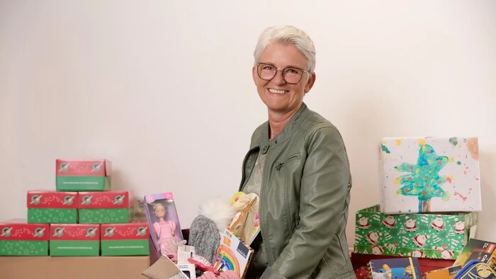 Eine Frau mit kurzen Haaren lächelt in die Kamera und hält Geschenke in der Hand.