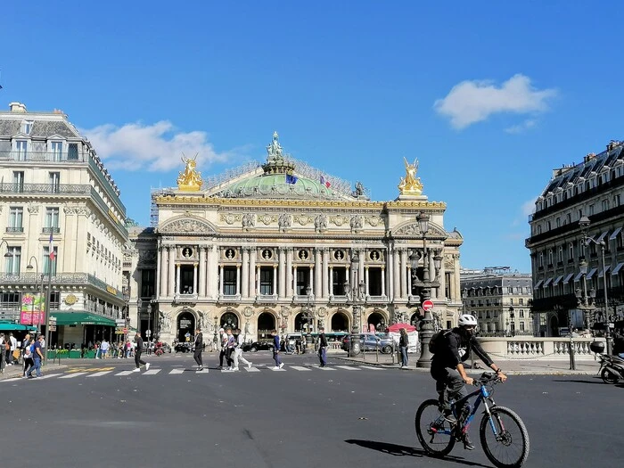 Bild zeigt die Opéra Garnier in Paris, davor Radfahrer und Fußgänger auf einer breiten Straße.