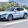 Ein hellblaues Auto mit der Aufschrift „Polizia“ fährt auf einer Straße, im Hintergrund sind Bäume
