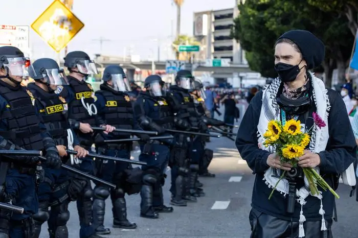 EDITORS NOTE: Graphic content / TOPSHOT - A demonstrator holding flowers walks past police officers as protesters clash with law enforcement in the streets surrounding the federal building during a protest following federal immigration operations in Los Angeles, California, on June 8, 2025. Demonstrators torched cars and scuffled with security forces in Los Angeles on June 8, as police kept protestors away from the National Guard troops President Donald Trump sent to the streets of the second biggest US city. (Photo by RINGO CHIU / AFP)