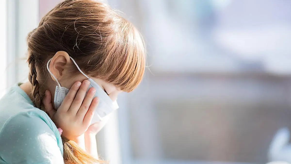 Adorable sad child girl in medical mask by the window. Quarantine. Coronavirus, COVID19. Epidemic. Closeup portrait, copy space
