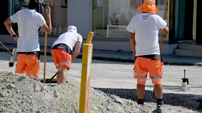 Man sieht drei Straßenarbeiter von hinten bei der Arbeit, zwei von ihnen wirken erschöpft