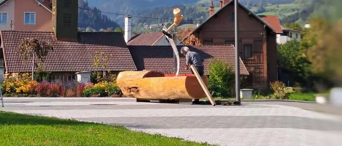 Ein Dorfplatz, im Hintergrund sieht man Häuser und Berge, im Fokus ist ein Brunnen aus Holz